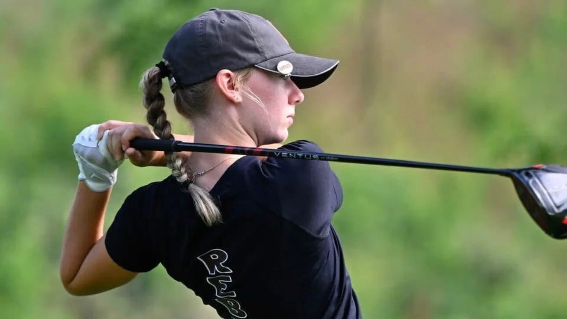 Ava Klips at the top of her follow-through after a tee shot, driver over her shoulder, trees behind her