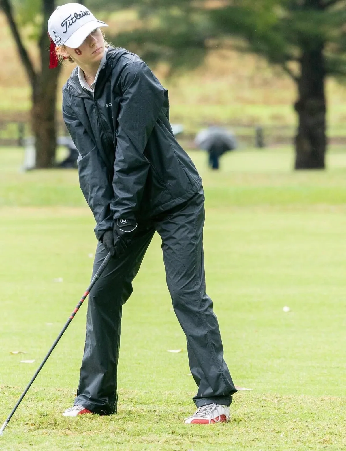 Ava Klips in black rain gear playing a pitch shot from a wet fairway